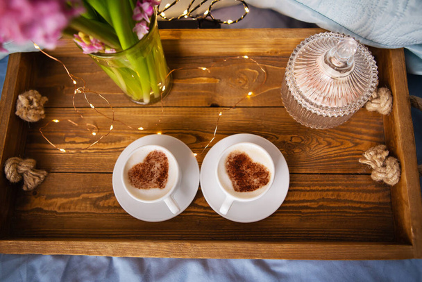 Two white cups of cappuccino stand on an old wooden tray. Garland, beautiful blinking. Top view, close-up. - Photo, Image