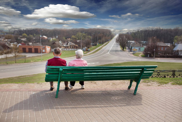 Starsi ludzie siedzą na zielonej ławce. Spogląda na odległość na drodze i piękne niebo z chmurami i lasem. - Zdjęcie, obraz