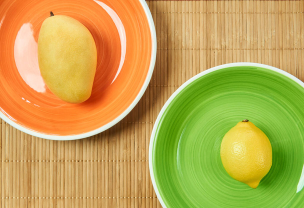 κίτρινα φρούτα - γλυκό μάνγκο σε ένα πορτοκαλί πιάτο και ξινό λεμόνι σε ένα πράσινο πιάτο - σε ένα στρώμα ζαχαροκάλαμου εξυπηρετούν - Φωτογραφία, εικόνα