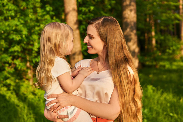 Улыбающаяся красивая молодая женщина нежно смотрит на свою маленькую дочь, которая держит на руках, на естественном фоне
 - Фото, изображение