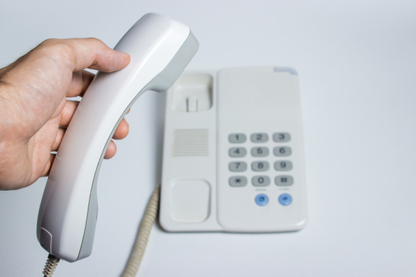 Рука держит телефонный приемник со стационарного телефона. Белый стационарный телефон с кнопками. Фото на белом фоне
 - Фото, изображение