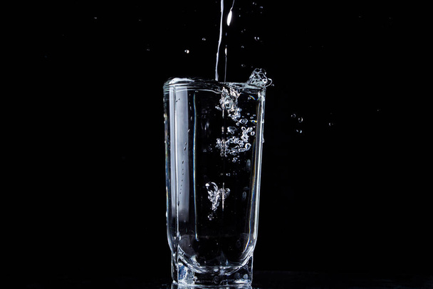 Egy vízfolyam ömlik egy pohár vízbe. A víz elárasztja az üveget. Fénykép fekete háttérrel. Tiszta és tisztított víz ömlött ki egy pohárból - Fotó, kép