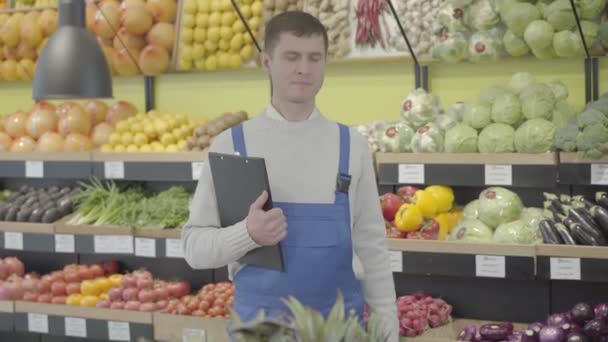 Young positive Caucasian employee posing in grocery with folder and eggplant. Portrait of smiling man in uniform working in supermarket. Business, commerce, profession, lifestyle. S-log 2. - Video, Çekim