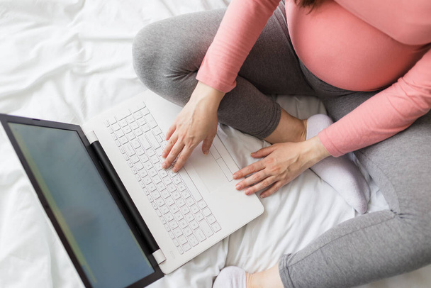 High angle view of young pregnant woman sitting on bed and using laptop. - Foto, Bild