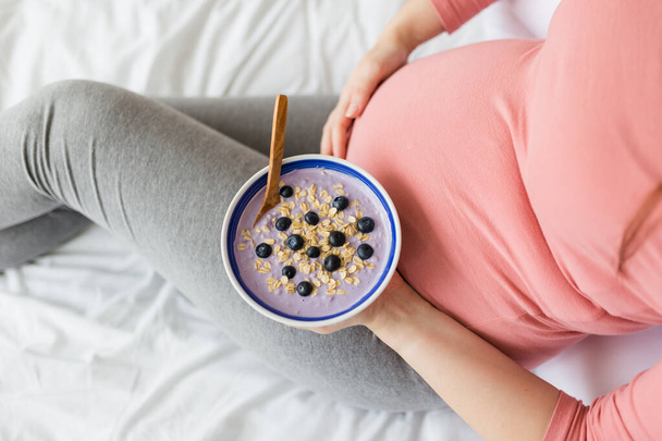 Female holding bowl with fresh muesli on bed. Pregnancy and healthy lifestyle concept. - Φωτογραφία, εικόνα