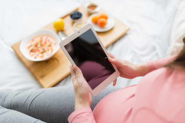 Close up of young pregnant woman using digital tablet next to fresh oatmeal and fruits on small table in bed. - Foto, Bild