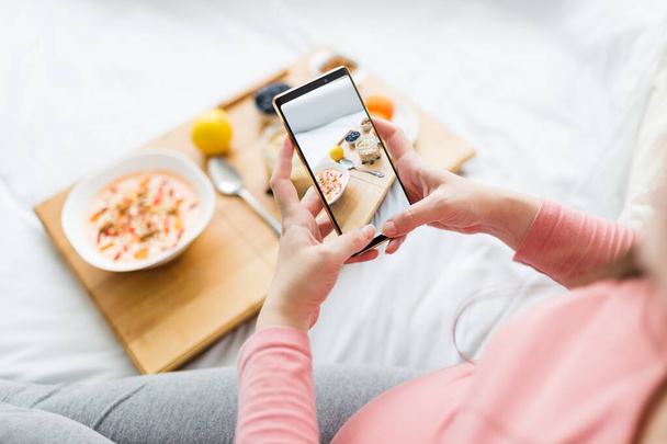 Close up of young pregnant woman taking photo of fresh oatmeal and fruits on small table in bed with smartphone. - Fotoğraf, Görsel
