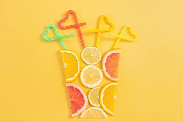 Kreatywna koncepcja soku cytrusowego, smoothie, letnich drinków. Kawałki pomarańczy, grejpfrutów, cytryny i limonki są składane w kształcie szkła na żółtym tle. Top - czerwone, żółte, zielone, pomarańczowe rurki. - Zdjęcie, obraz