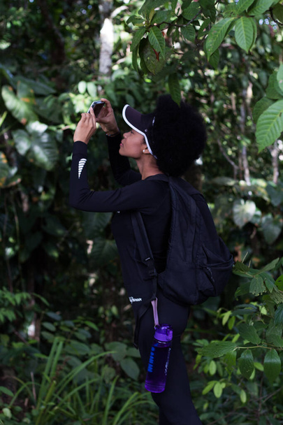 femme noire vêtue de noir prenant des photos dans la forêt avec un téléphone cellulaire bleu - Photo, image