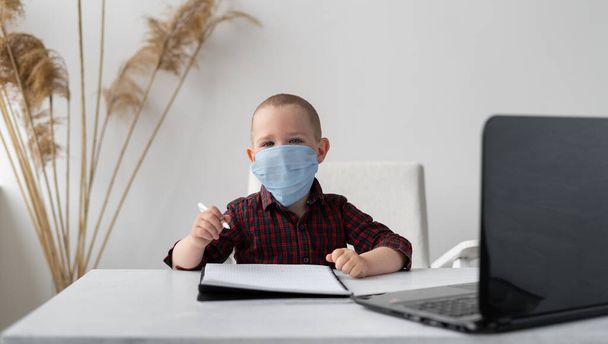 Boy schoolboy in a medical mask does homework online. In front of him is a laptop. There is a notebook on the table. Distance Learning and Quarantine. Self-study at home on the Internet. Isolation of schoolchildren. Place for text. - Photo, Image