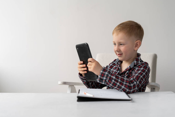 Schoolboy blond sits on a chair in front of the table. Distance Learning and Quarantine. The child has a tablet in his hands and a notebook for writing on the table. He is reading homework and smiling. Online education on the Internet for students. P - 写真・画像