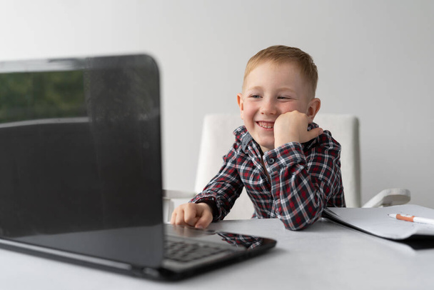 A schoolboy is sitting at home at a laptop and smiling. Online lessons. Education of children in quarantine. Homework on the webcam. Distance learning and lifestyle. Self-isolation of a schoolboy. A place for text. - Photo, Image