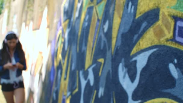 Jolie fille caucasienne avec écouteurs et smartphone passant devant la caméra. Extérieur, mur avec graffiti en arrière-plan. 4k, mouvement lent 75fps - Séquence, vidéo