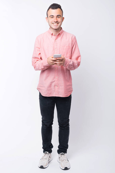Leveä hymyilevä nuori mies katsoo kameraa tekstaillessaan puhelimessa valkoisella taustalla - Valokuva, kuva