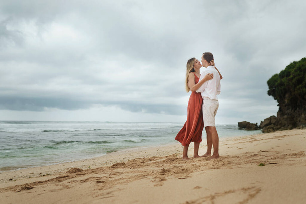 Beautiful young couple in love is hugging on the beach by the ocean on a background of cloudy gray sky, rocks and waves. Girl with blond hair in a red dress and a guy in a white shirt and shorts - Фото, изображение