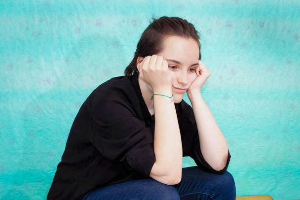 Une adolescente en chemise noire est assise avec un visage affligé soutenu avec ses joues. Plan moyen, fond turquoise. - Photo, image