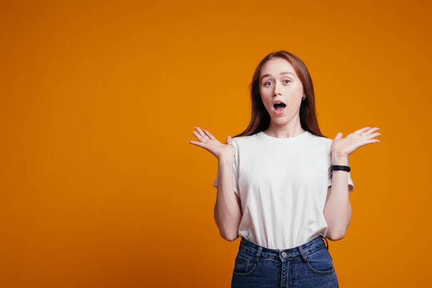 Portrait of a red-haired European girl who spreads her arms to the sides and looks to the side, not knowing what to choose. Orange background. - Valokuva, kuva