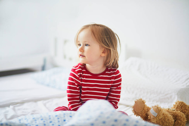 Happy toddler girl in striped red and white pajamas sitting on bed right after awaking. Day naps for small kids - Foto, Imagen