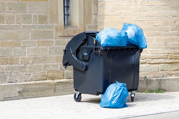 μαύρο πλαστικό κάδο με πλήρη μπλε σακούλες σκουπιδιών στο δρόμο - Φωτογραφία, εικόνα