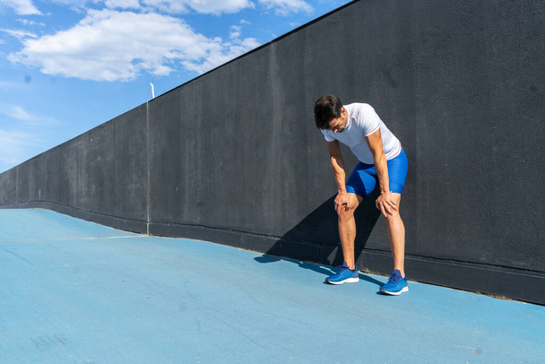 Hombre joven corriendo en pantalones azules y camisa blanca descansando y recuperándose en la pared negra a pleno sol. Concepto de motivación, carrera
 - Foto, imagen