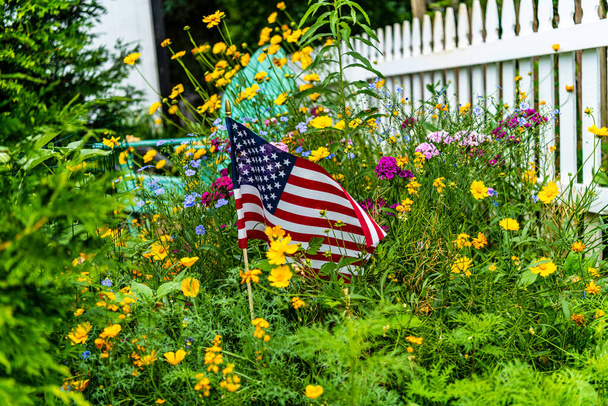 Горизонтальный открытый американский флаг в саду из диких цветов с белым забором для пикета и синим задним сиденьем стула adirondack
 - Фото, изображение