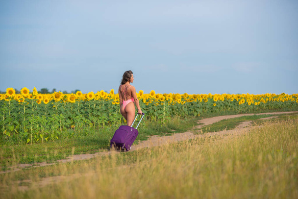 Beautiful woman in a pink swimsuit travels through the countryside with a suitcase. A girl in a monokini is walking alone on a field of sunflowers with a big bag. Hiking. Rest and social distance. - Photo, Image