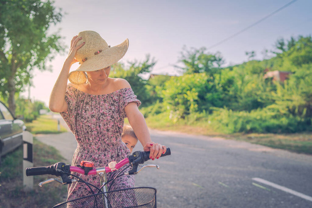 Jonge vrouw met stuur grip duwen fiets in een zomer zonnige dag dragen jurk en hoed in de openlucht asfalt - Foto, afbeelding