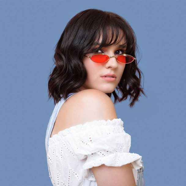 Lovely brunette woman with orange glasses looking back to camera and posing on a blue studio wall - Photo, Image