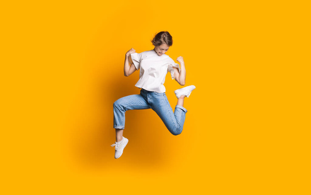 Hüpfende Kaukasierin in Jeans und weißem T-Shirt lächelt und wirbt auf einer gelben Studiowand für etwas - Foto, Bild