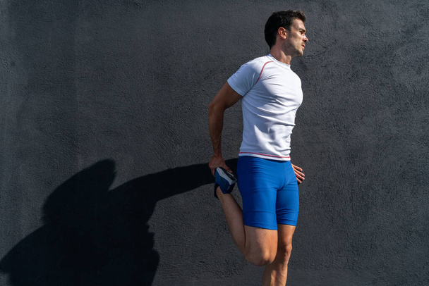 Fiatal férfi fut kék nadrágban és fehér ing pihen és lábadozik a fekete falon teljes napsütésben, csinál láb nyújtás. Motivációs koncepció, karrier - Fotó, kép