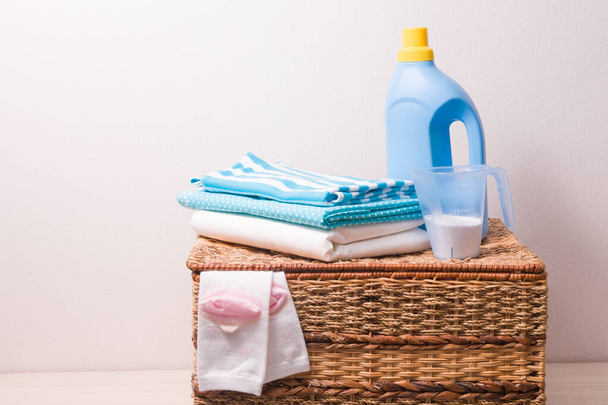 clothes in light blue tones, strange powder in a measuring cup and gel for washing colored clothes in a blue plastic bottle on a wicker laundry basket, children's socks stick out of the baske - Fotoğraf, Görsel