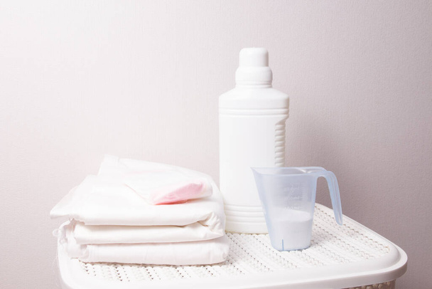 the concept of washing white linen, bleach and detergent on a white laundry basket, a stack of white linen and baby socks, light background, copy space - Photo, Image