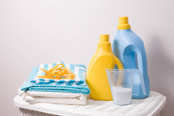 a stack of colored laundry, clothes pegs, laundry detergent in a measuring cup and washing gels in yellow and blue bottles on a white plastic basket for dirty laundry, copy space - Photo, Image