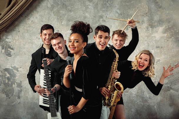 Cheerful international music group on a gray wall background, a group of musicians posing on camera in the hands of various instruments, guitars, saxophone. Copy space. - Photo, Image