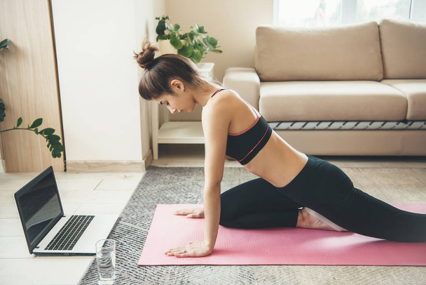 Side view photo of a caucasian woman doing fitness at home online using a laptop and glass with water on floor wearing sportswear - Фото, зображення