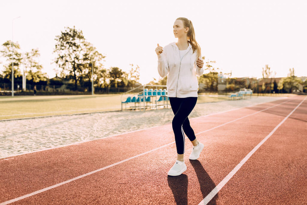 Athletic young woman in sportswear running around the stadium at dawn - Фото, изображение