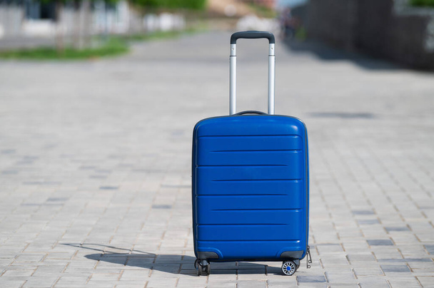 Blue suitcase on wheels on the sidewalk. Luggage is on the street of the city and ready for travel. Large bag for moving. No people. - 写真・画像