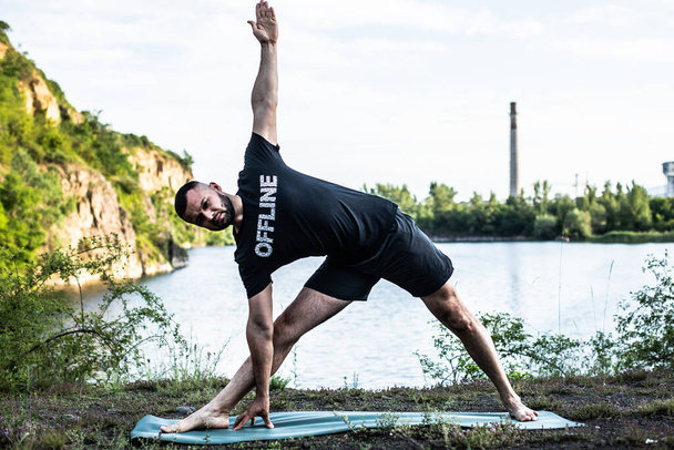 Hombre inspirado haciendo asanas de yoga en el lago de la cantera. Ciudadano joven haciendo ejercicio al aire libre y de pie en postura de ángulo lateral de yoga. Fitness al aire libre y concepto de equilibrio de vida. - Foto, Imagen