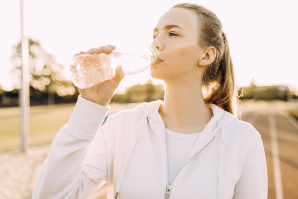 Atletik güzel koşucu koştuktan sonra plastik şişeden su içer. Bir kız açık hava fitness derslerinden sonra susuzluğunu giderir. - Fotoğraf, Görsel