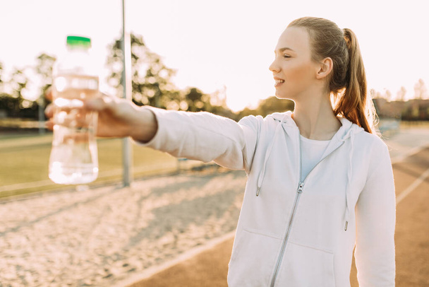 Beautiful athletic female runner standing outdoors at the stadium holding a water bottle. Fitness woman takes a break during a workout - Fotoğraf, Görsel