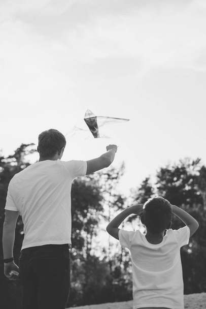 Vater und Sohn stehen mit dem Rücken im Rahmen auf einem Hügel und lassen einen Drachen steigen. Schwarz-Weiß-Fotografie. - Foto, Bild