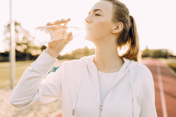 Athletic beautiful runner drinks water from a plastic bottle after running. A girl quenches her thirst after outdoor fitness classes - Fotoğraf, Görsel