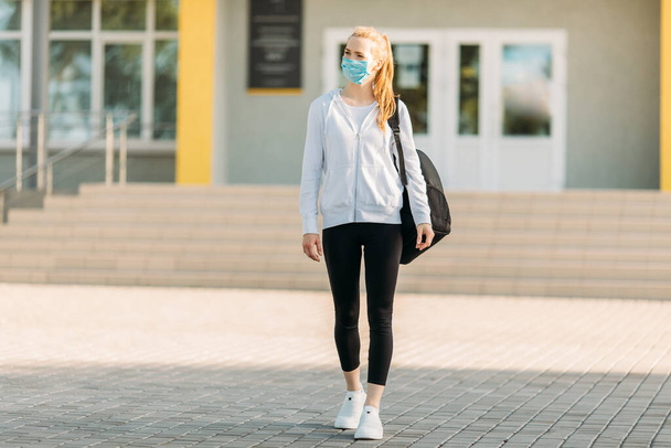 a young girl in a medical protective mask, standing with a briefcase against the background of a building, in the open air. Quarantine, coronavirus - Φωτογραφία, εικόνα