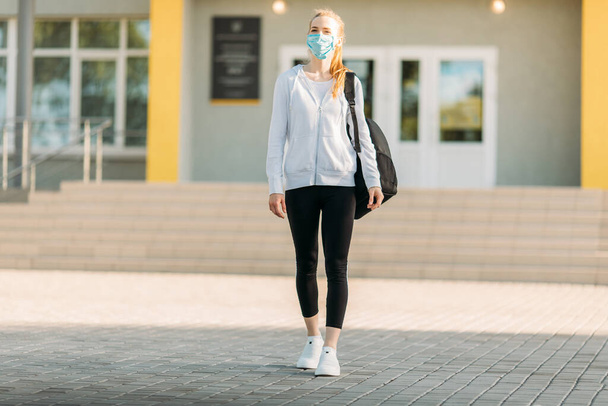 a young girl in a medical protective mask, standing with a briefcase against the background of a building, in the open air. Quarantine, coronavirus - Zdjęcie, obraz