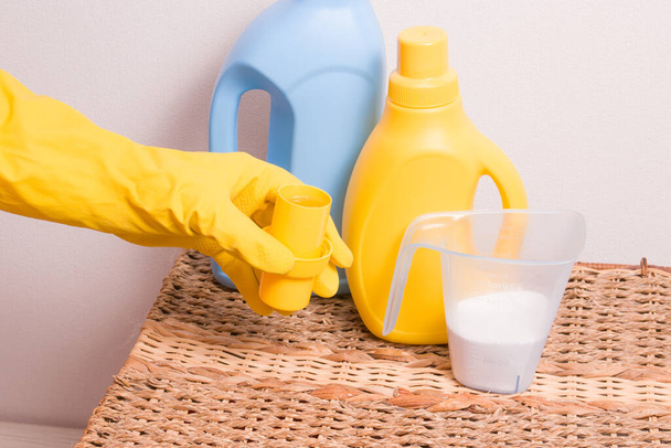 a female hand in a yellow rubber glove holds a yellow measuring cap with washing gel, washing powder, gel and fabric softener in bottles without labels on a wicker basket - Фото, зображення