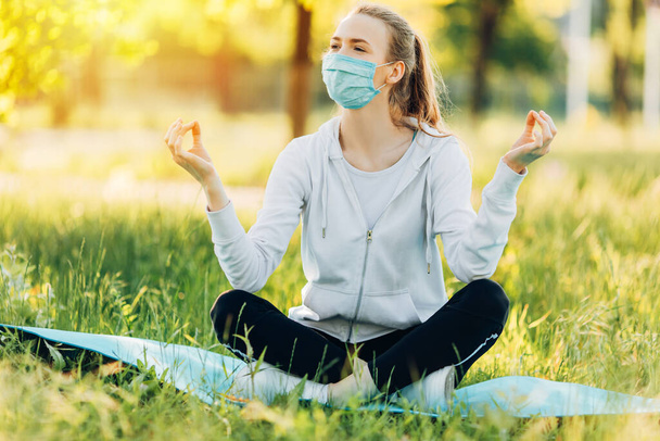 menina em uma máscara protetora médica medita na posição de lótus na grama verde ao amanhecer. Estilo de vida saudável e relaxamento
. - Foto, Imagem