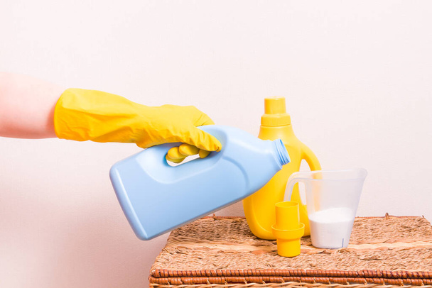 woman in yellow rubber gloves pours a softener for kids clothes in little yellow cup for washing liquid - Foto, Imagen