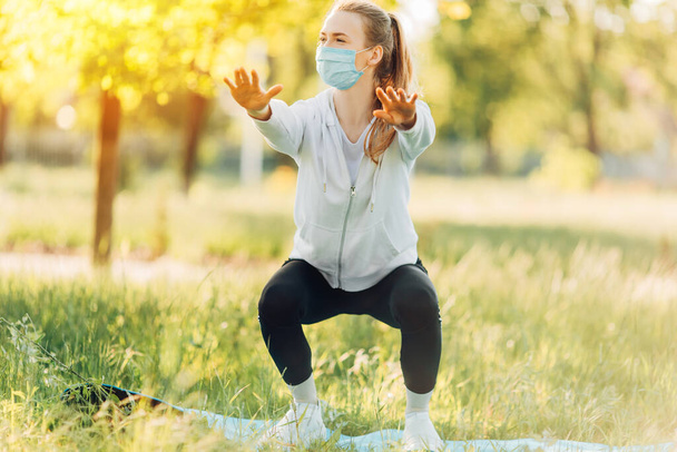 Athletic young woman in a medical protective mask, doing sports in the Park in the morning, women's training on a yoga Mat - Foto, Imagen