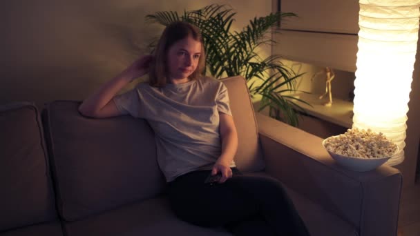 Girl sitting on the couch switches channels on the TV. Behind home plant - Πλάνα, βίντεο