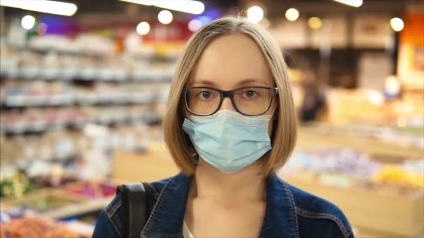 Ritratto di una giovane ragazza caucasica in maschera protettiva medica e occhiali. Supermercato al rallentatore, primo piano. - Filmati, video